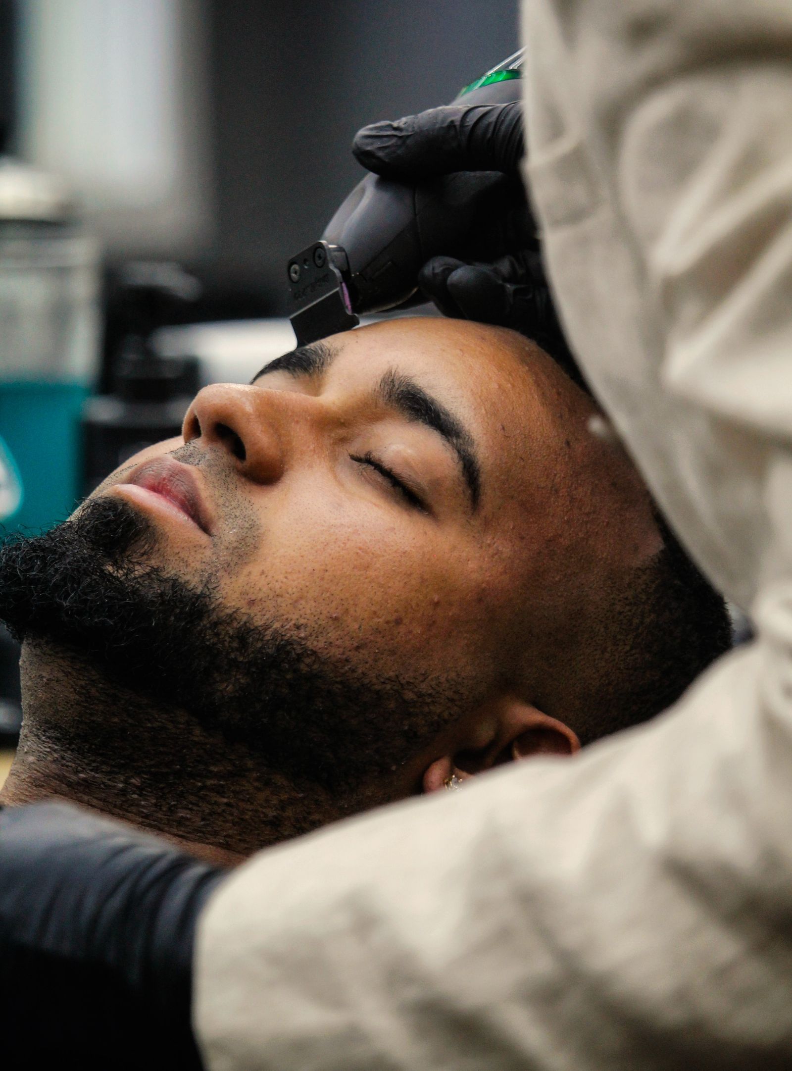 A reclined client with eyes closed while a gloved barber lines up his hairline.