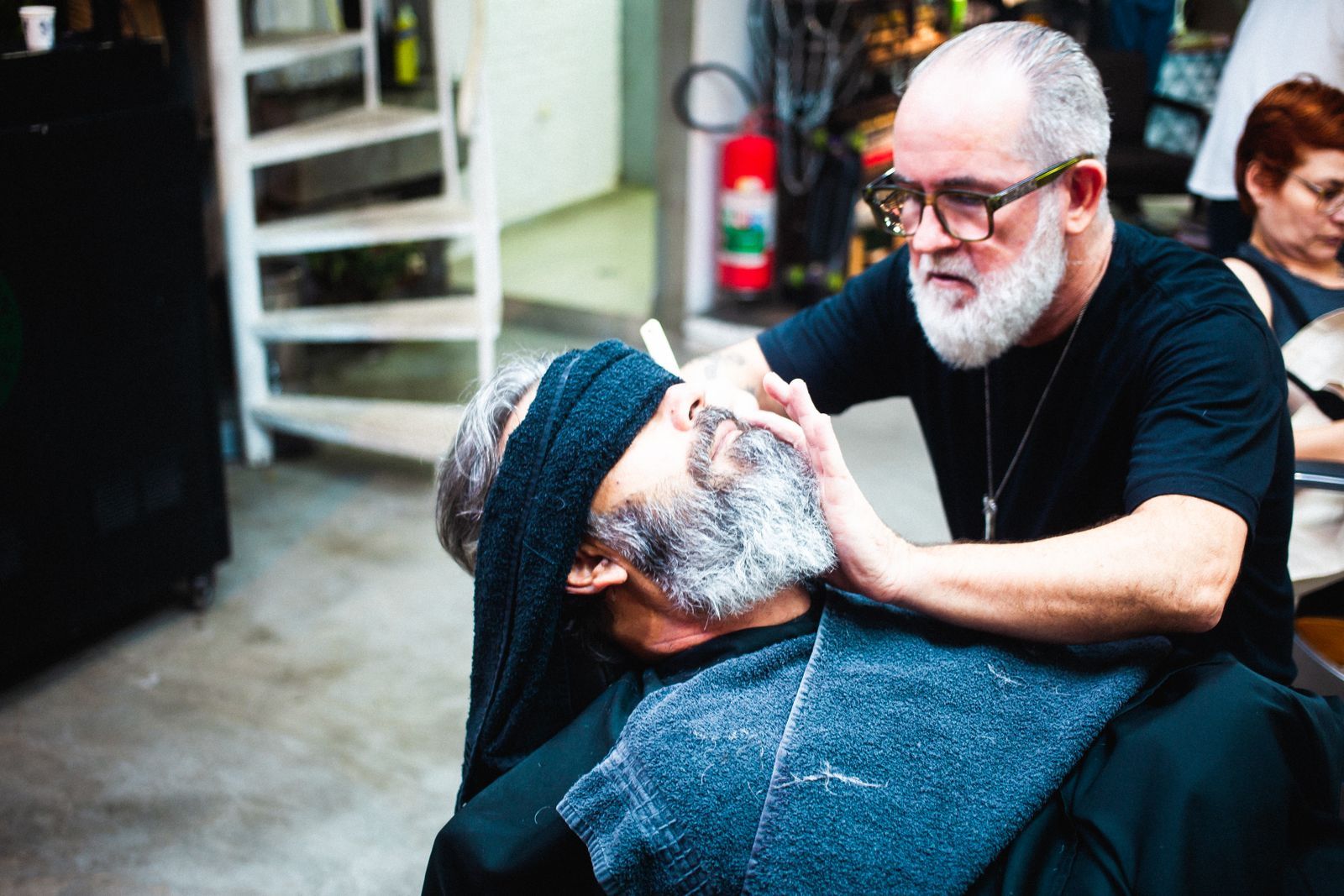 A barber with a white beard trims a client's grey beard as he leans back under a towel.